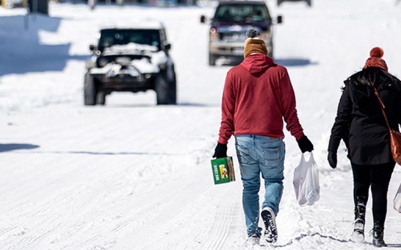 two people walk through snow carrying groceries as vehicles approach in the other direction